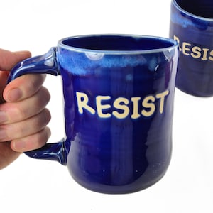 May include: A cobalt blue ceramic mug with the word "RESIST" in cream-colored letters. The mug has a large handle and a slightly flared rim. Another mug with the word "RESIST" is visible in the background.