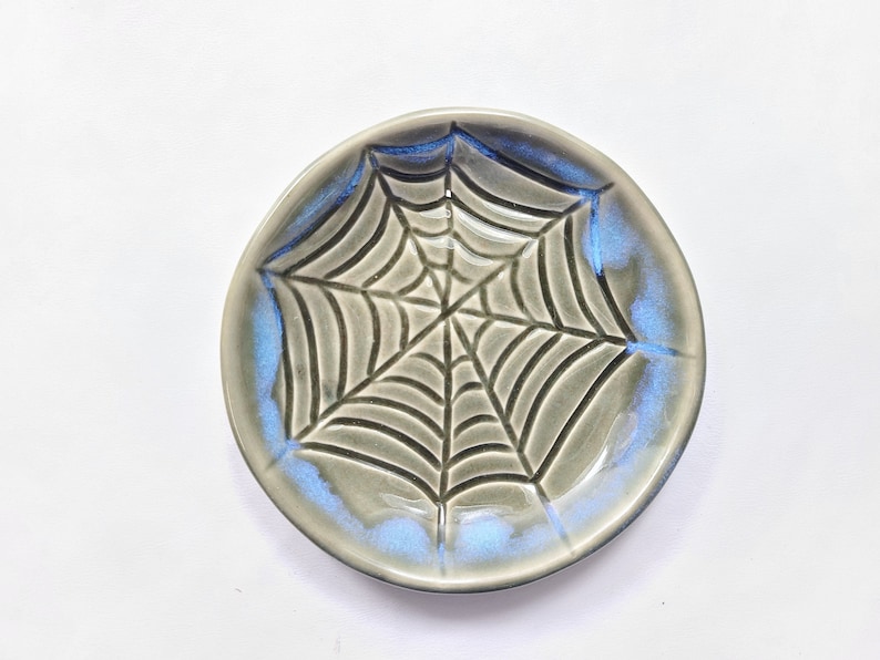 May include: A small, round ceramic dish with a spiderweb design. The dish is gray with a blue iridescent glaze around the rim. The spiderweb is etched into the surface.