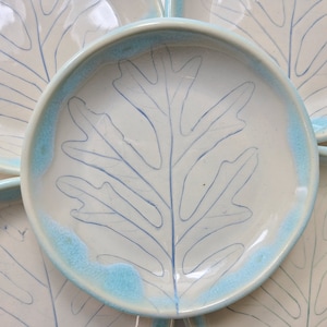 May include: Set of four ceramic plates with a light blue rim and a white center. Each plate features a blue outline of an oak leaf.