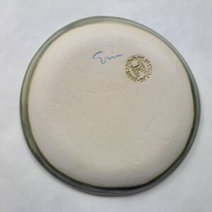 May include: A handmade ceramic plate with a light gray rim and a creamy white interior. The bottom of the plate has blue script and a green stamp that reads "MADE IN ITALY". The plate is a unique piece of pottery.