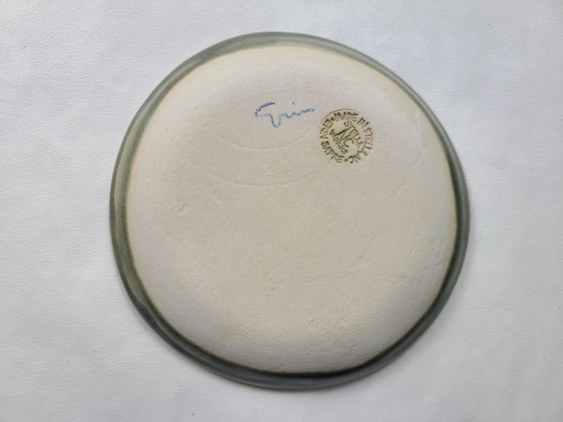 May include: A handmade ceramic plate with a light gray rim and a creamy white interior. The bottom of the plate has blue script and a green stamp that reads "MADE IN ITALY". The plate is a unique piece of pottery.