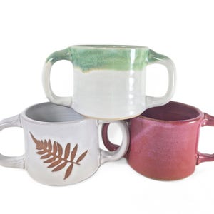 May include: Three ceramic mugs with unique designs. One mug is white with a brown fern, another is white with a green top, and the third is red. Each mug has two handles.