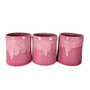 May include: Three cylindrical ceramic cups featuring a vibrant magenta glaze with a lighter pink drip effect cascading from the top rim. Each cup has a smooth, glossy finish and a consistent, simple shape without handles, arranged in a horizontal row.