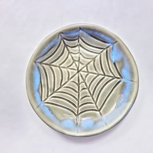 May include: A ceramic plate with a spiderweb design. The plate is round with a gray base and a blue iridescent glaze around the rim. The spiderweb is etched into the plate's surface.