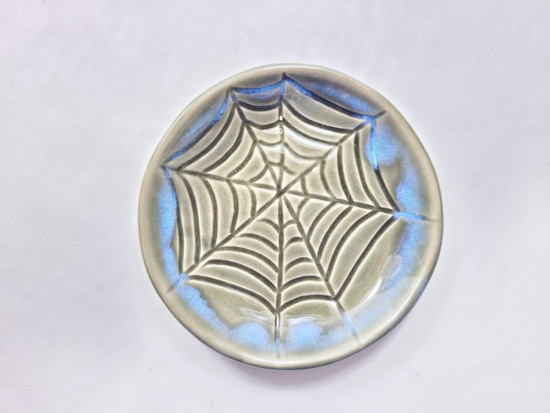 May include: A ceramic plate with a spiderweb design. The plate is round with a gray base and a blue iridescent glaze around the rim. The spiderweb is etched into the plate's surface.