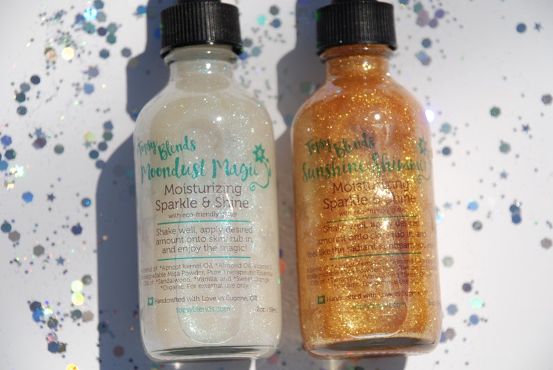 May include: Two glass bottles with gold and white glitter inside. The bottles are labeled "Topsy Blends Moondust Magic Moisturizing Sparkle & Shine" and "Topsy Blends Sunshine Shimmer Moisturizing Sparkle & Shine".