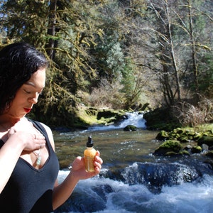 May include: A woman wearing a black tank top stands in a forest setting with a small bottle of liquid in her hand. The bottle is clear with a gold cap and a dropper. The woman is looking down at the bottle. The background is a lush forest with a river flowing through it.