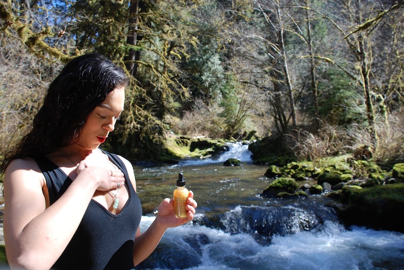 May include: A woman wearing a black tank top stands in a forest setting with a small bottle of liquid in her hand. The bottle is clear with a gold cap and a dropper. The woman is looking down at the bottle. The background is a lush forest with a river flowing through it.