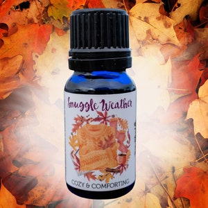 May include: A small blue glass bottle with a black cap and a white label that says "Snuggle Weather" with a picture of a sweater, a mug, and cinnamon sticks. The label also says "Cozy & Comforting".