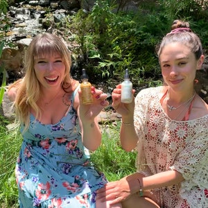 May include: Two people are sitting outdoors in a grassy area with a small stream behind them. They are both holding small glass bottles with droppers. The bottle on the left is gold and the bottle on the right is white.