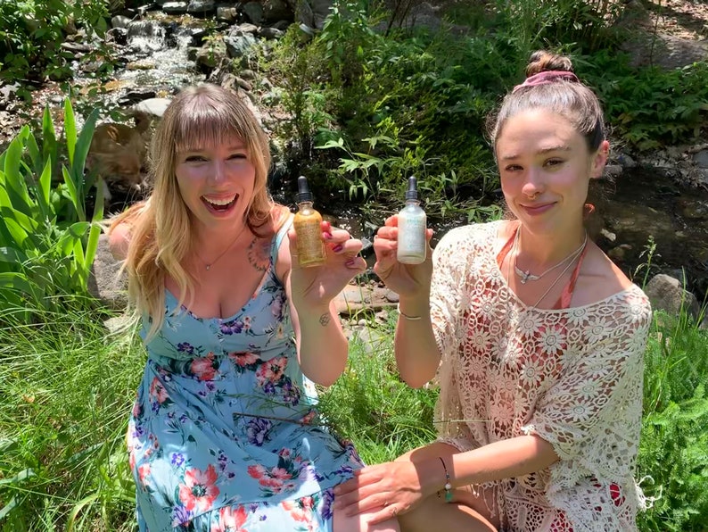 May include: Two people are sitting outdoors in a grassy area with a small stream behind them. They are both holding small glass bottles with droppers. The bottle on the left is gold and the bottle on the right is white.
