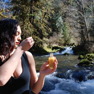 May include: A woman in a black tank top holds a small, clear glass bottle with a gold liquid inside. The bottle is in front of a rushing river with rocks and green moss.