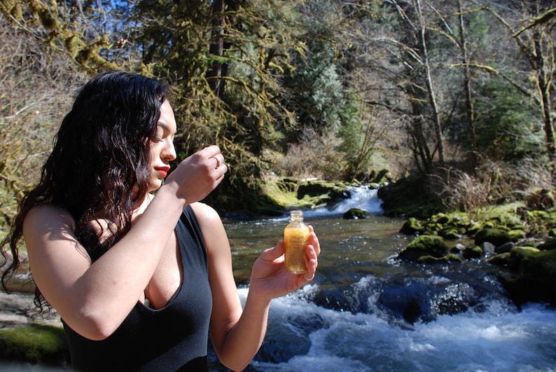 May include: A woman in a black tank top holds a small, clear glass bottle with a gold liquid inside. The bottle is in front of a rushing river with rocks and green moss.
