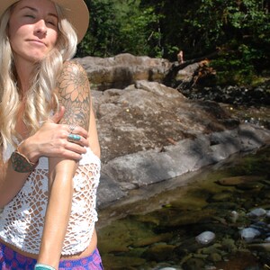 May include: A woman with long blonde hair wearing a white crochet top, a wide-brimmed straw hat, and a floral tattoo on her arm. She is standing in a stream with rocks and water.