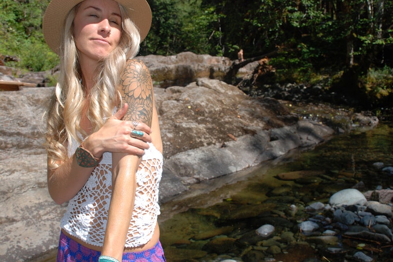 May include: A woman with long blonde hair wearing a white crochet top, a wide-brimmed straw hat, and a floral tattoo on her arm. She is standing in a stream with rocks and water.