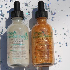 May include: Two glass bottles with dropper tops filled with shimmering body oil. The bottles are labeled "Moondust Magic" and "Sunshine Shimmer" with the text "Moisturizing Sparkle & Shine" on each. The bottles are on a white background with iridescent glitter.