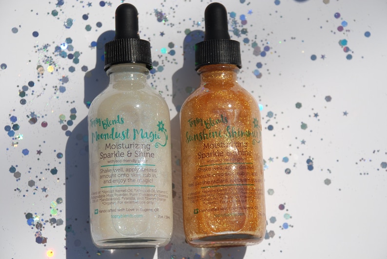 May include: Two glass bottles with dropper tops filled with shimmering body oil. The bottles are labeled "Moondust Magic" and "Sunshine Shimmer" with the text "Moisturizing Sparkle & Shine" on each. The bottles are on a white background with iridescent glitter.