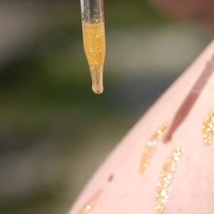 May include: A clear glass dropper bottle with a gold liquid inside. The liquid is being dripped onto a person's arm, leaving a trail of gold glitter.
