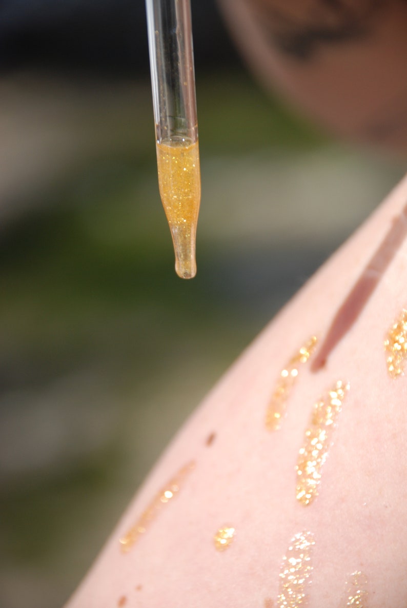May include: A clear glass dropper bottle with a gold liquid inside. The liquid is being dripped onto a person's arm, leaving a trail of gold glitter.