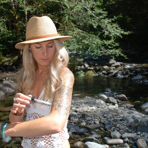 May include: A woman wearing a straw hat and a white crocheted top applies a clear liquid from a small bottle to her arm. She is standing in a forest setting near a stream.