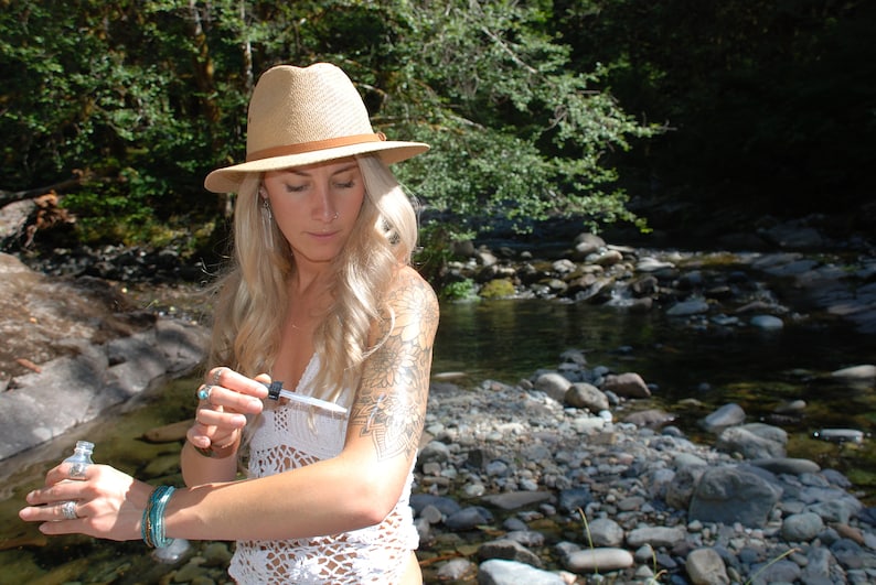 May include: A woman wearing a straw hat and a white crocheted top applies a clear liquid from a small bottle to her arm. She is standing in a forest setting near a stream.