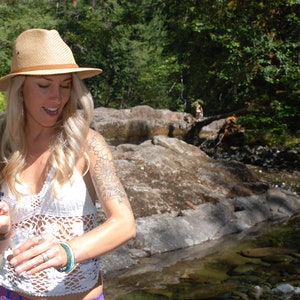 May include: A woman wearing a white crochet top and a straw hat with a brown band. She is standing in a forest setting with a small stream in the background. She has a tattoo on her arm and is holding a small bottle in her hand.