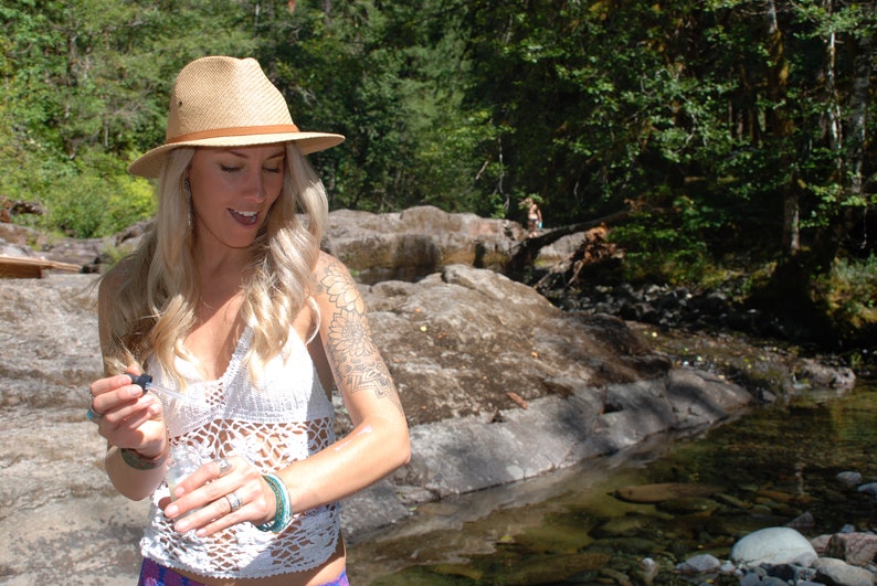 May include: A woman wearing a white crochet top and a straw hat with a brown band. She is standing in a forest setting with a small stream in the background. She has a tattoo on her arm and is holding a small bottle in her hand.