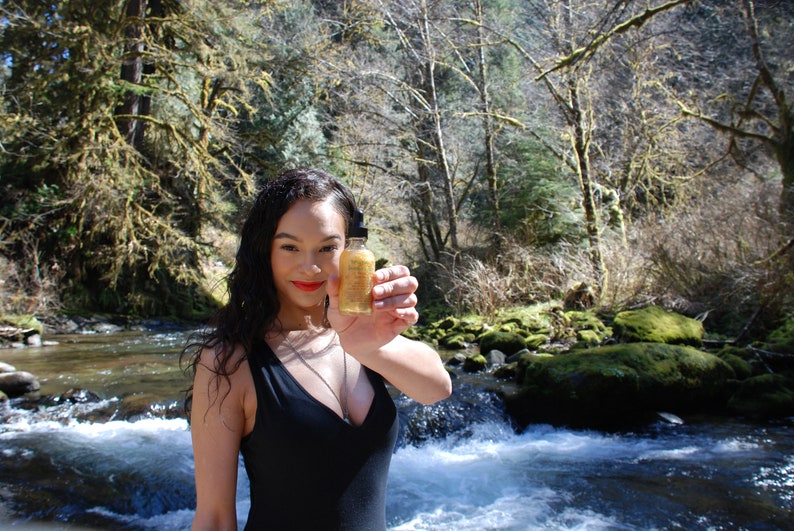 May include: A woman in a black swimsuit stands in a river, holding a small glass bottle with a gold label. The bottle is filled with a clear liquid. The river is surrounded by trees and rocks.