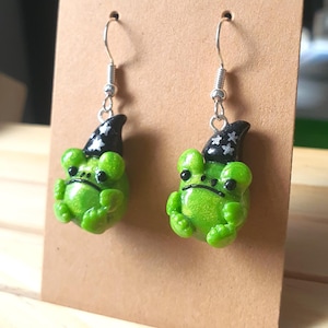 May include: Handmade green frog earrings, each with a black wizard hat featuring white stars. The earrings are displayed on a brown card. These quirky earrings are a unique accessory, showcasing the craftsmanship and whimsical design.