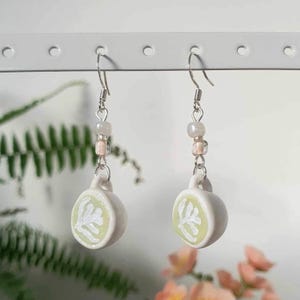 May include: A pair of white ceramic earrings shaped like teacups with a green leaf design. The earrings are strung on silver chains with small white and pink beads.