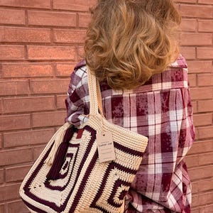 以下が含まれることがあります： ベージュとバーガンディ色の幾何学模様の編み物トートバッグ。バッグは長方形で、お揃いのハンドルと「Lunar cat」と書かれたタグが付いています。バッグは人の肩に掛かっています。