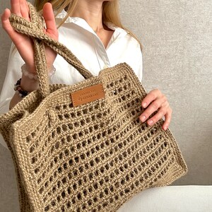 May include: A large, crocheted tote bag made of natural jute fibre. The bag has a rectangular shape with a wide, open top and a brown leather label that reads "CROCHET BAG LUNAR CAT".