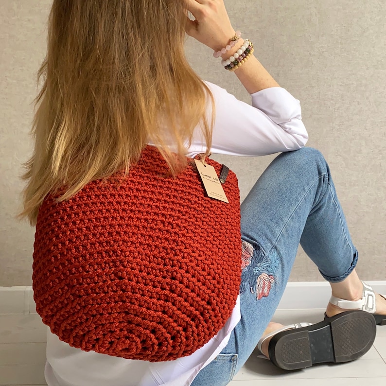May include: A rust-colored crocheted tote bag with a brown leather handle and a small brown tag that says "Lumber Life" hanging from the handle.