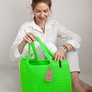 May include: A bright green crocheted tote bag with leather handles and a brown paper tag. The bag is sitting on a white surface.