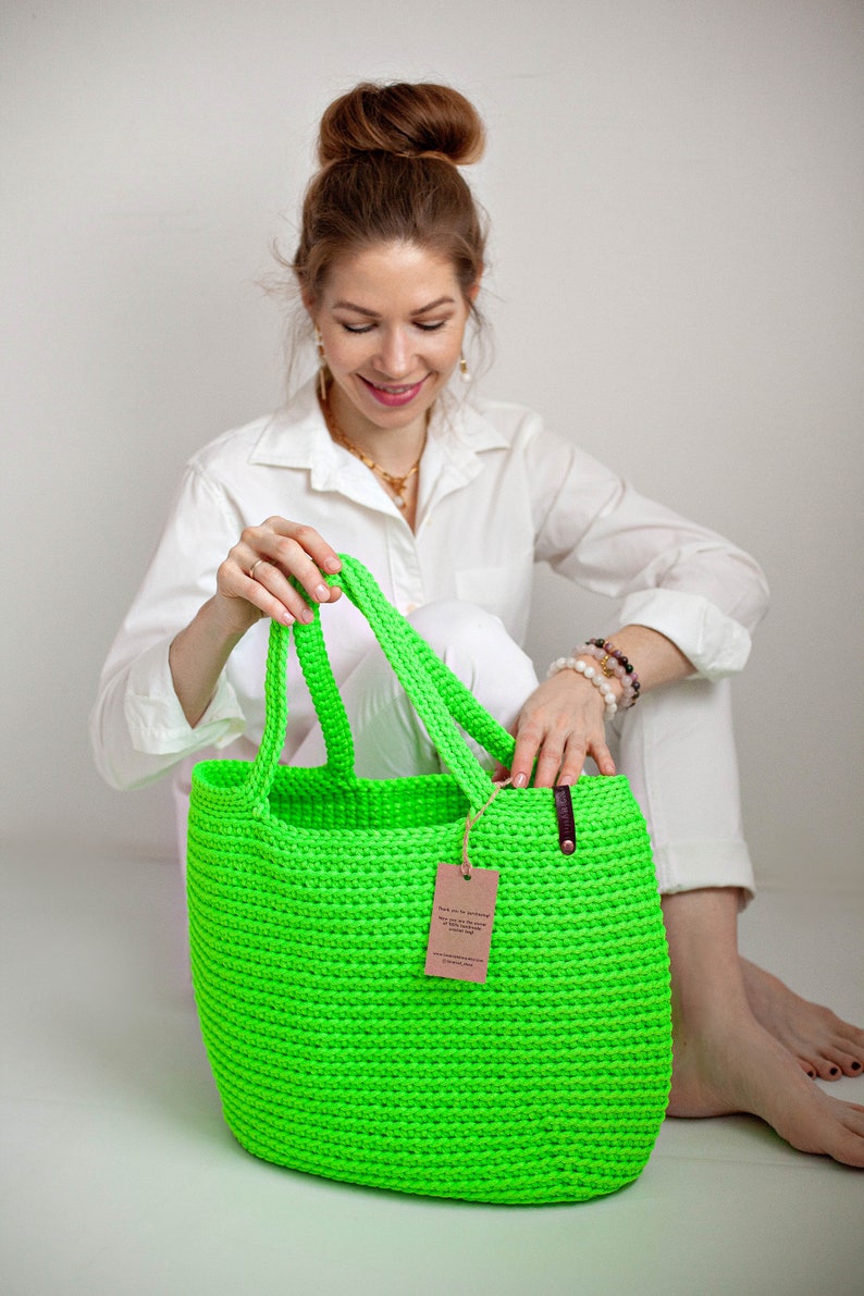 May include: A bright green crocheted tote bag with leather handles and a brown paper tag. The bag is sitting on a white surface.