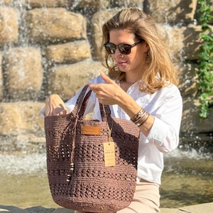 K&ouml;nnte beinhalten: Eine braune geh&auml;kelte Tragetasche mit einem Lederanh&auml;nger, auf dem "Handmade with Love" steht. Die Tasche wird von einer Person gehalten, die ein wei&szlig;es Hemd, eine Sonnenbrille und einen beigefarbenen Rock tr&auml;gt.