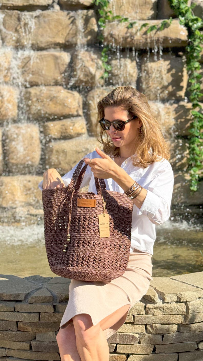 K&ouml;nnte beinhalten: Eine braune geh&auml;kelte Tragetasche mit einem Lederanh&auml;nger, auf dem "Handmade with Love" steht. Die Tasche wird von einer Person gehalten, die ein wei&szlig;es Hemd, eine Sonnenbrille und einen beigefarbenen Rock tr&auml;gt.