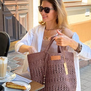 K&ouml;nnte beinhalten: Eine Frau mit Sonnenbrille und einem wei&szlig;en Hemd h&auml;lt eine braune geh&auml;kelte Tragetasche mit einem Lederanh&auml;nger, auf dem "Crochet by Handmade Co." steht.