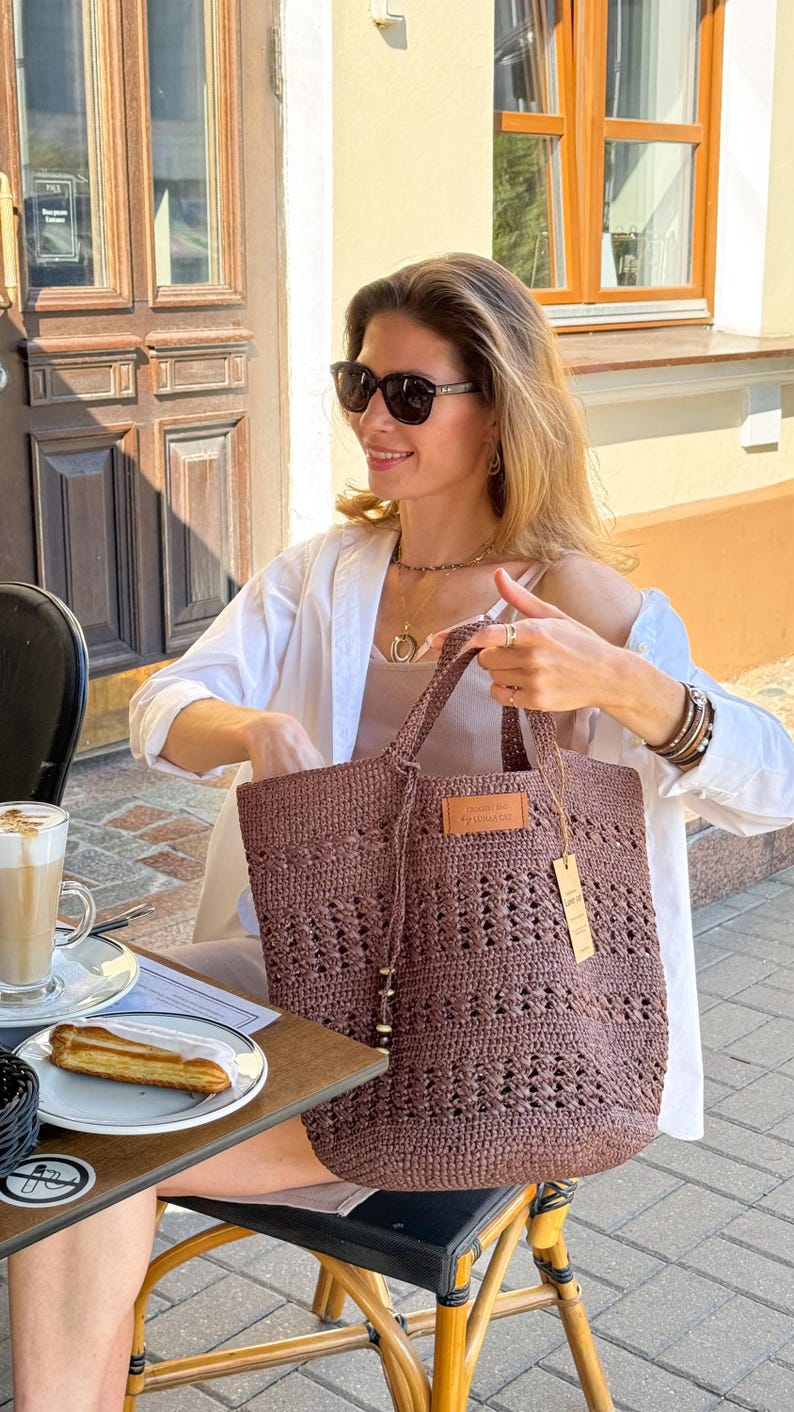 K&ouml;nnte beinhalten: Eine Frau mit Sonnenbrille und einem wei&szlig;en Hemd h&auml;lt eine braune geh&auml;kelte Tragetasche mit einem Lederanh&auml;nger, auf dem "Crochet by Handmade Co." steht.