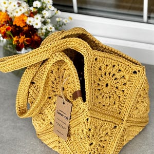 May include: Mustard yellow crochet tote bag with a square pattern. The bag has two handles and a tag that reads "Lunar cat". The bag is sitting on a gray surface with a vase of flowers in the background.