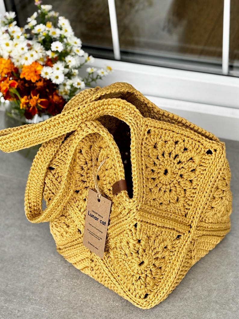 May include: Mustard yellow crochet tote bag with a square pattern. The bag has two handles and a tag that reads "Lunar cat". The bag is sitting on a gray surface with a vase of flowers in the background.