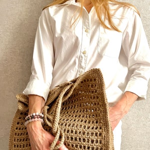 May include: A large, brown, crocheted tote bag with a woven pattern. The bag has a wide, flat base and two long, knotted handles.