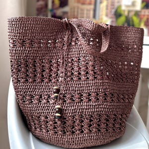 K&ouml;nnte beinhalten: Eine braune geh&auml;kelte Tragetasche mit einem gewebten Muster und Holzperlen-Akzenten. Die Tasche liegt auf einer wei&szlig;en Oberfl&auml;che.