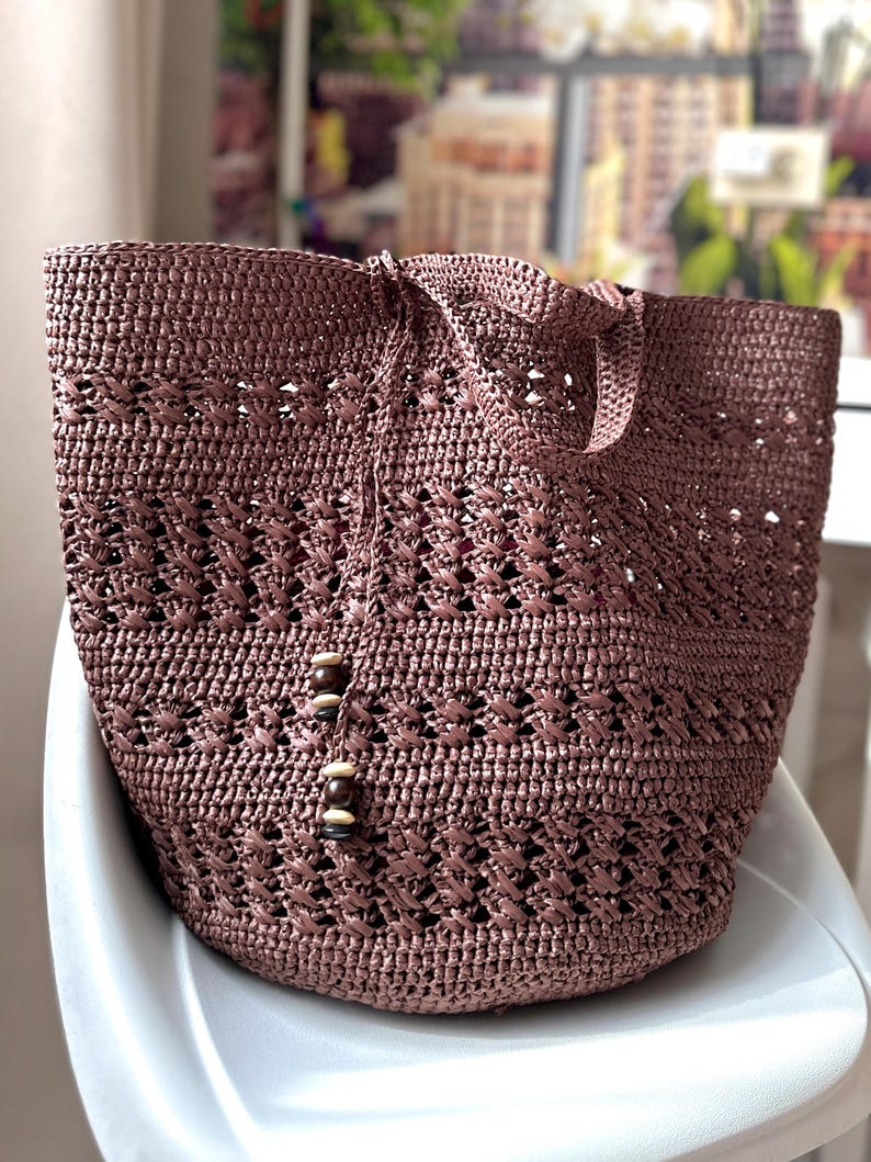 K&ouml;nnte beinhalten: Eine braune geh&auml;kelte Tragetasche mit einem gewebten Muster und Holzperlen-Akzenten. Die Tasche liegt auf einer wei&szlig;en Oberfl&auml;che.