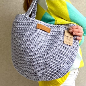 May include: A lavender crocheted tote bag with a brown leather tag that says "Crochet bag by LunarCat". The bag is being held by a person wearing a yellow and green sweater.