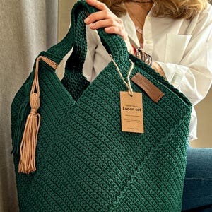 May include: A handmade, dark green crochet tote bag with a geometric design. The bag features a matching handle and a decorative tassel with tan accents. A small tag with the text "Lunar cat" is attached.