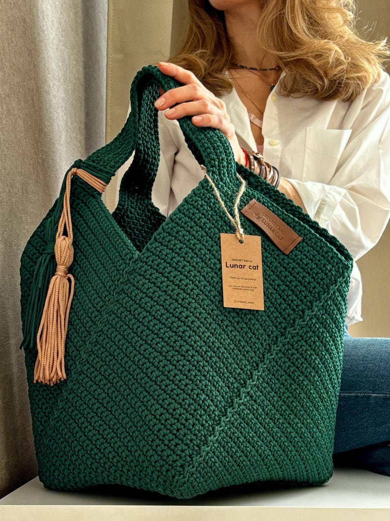 May include: A handmade, dark green crochet tote bag with a geometric design. The bag features a matching handle and a decorative tassel with tan accents. A small tag with the text "Lunar cat" is attached.