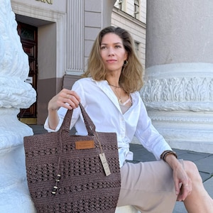 K&ouml;nnte beinhalten: Eine Frau sitzt auf einer Treppe und h&auml;lt eine braune geh&auml;kelte Tragetasche. Die Tasche hat ein Leder-Etikett mit dem Wort "Handmade". Die Frau tr&auml;gt ein wei&szlig;es Hemd und einen beigefarbenen Rock.