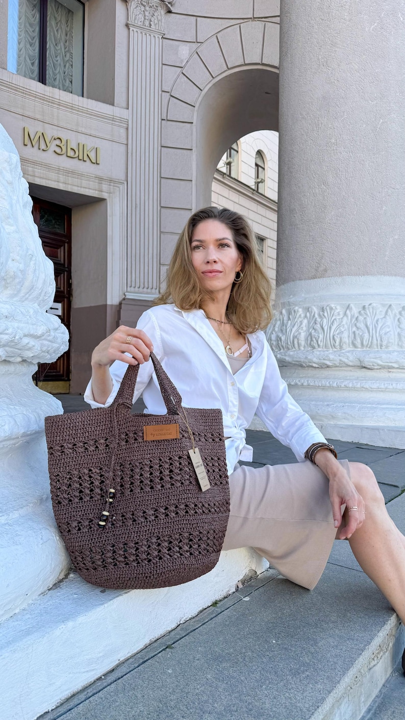 K&ouml;nnte beinhalten: Eine Frau sitzt auf einer Treppe und h&auml;lt eine braune geh&auml;kelte Tragetasche. Die Tasche hat ein Leder-Etikett mit dem Wort "Handmade". Die Frau tr&auml;gt ein wei&szlig;es Hemd und einen beigefarbenen Rock.