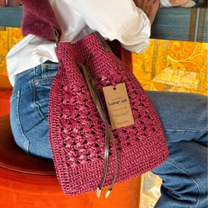 May include: A burgundy crochet bucket bag with brown leather straps and a brown paper tag that reads "Crochet bag by Lunar cat".
